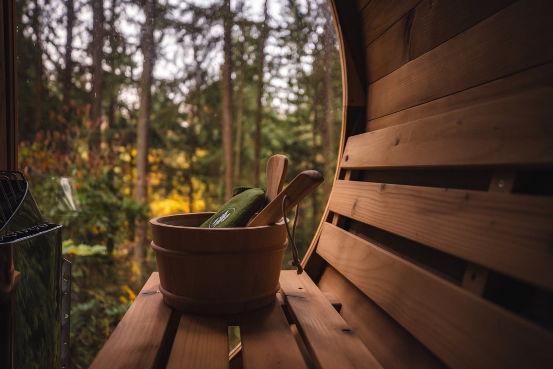 A sunlit sauna interior seen through the open glass door, with a wooden bucket and ladle on the bench