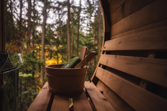 A sunlit sauna interior seen through the open glass door, with a wooden bucket and ladle on the bench