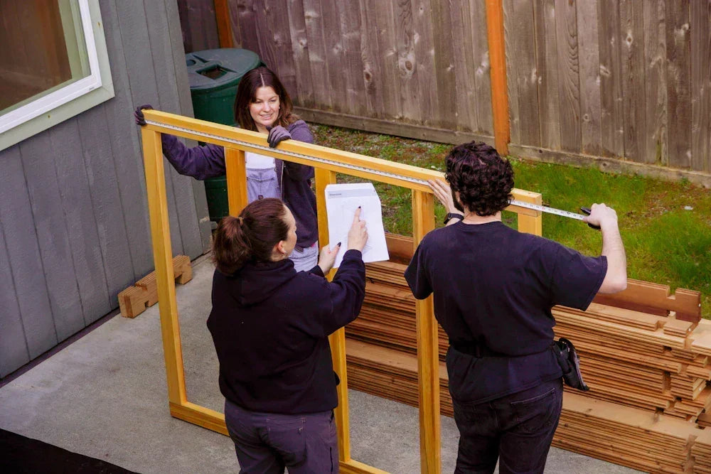 Three people holding up a wooden sauna wall frame during assembly, with stacked lumber nearby