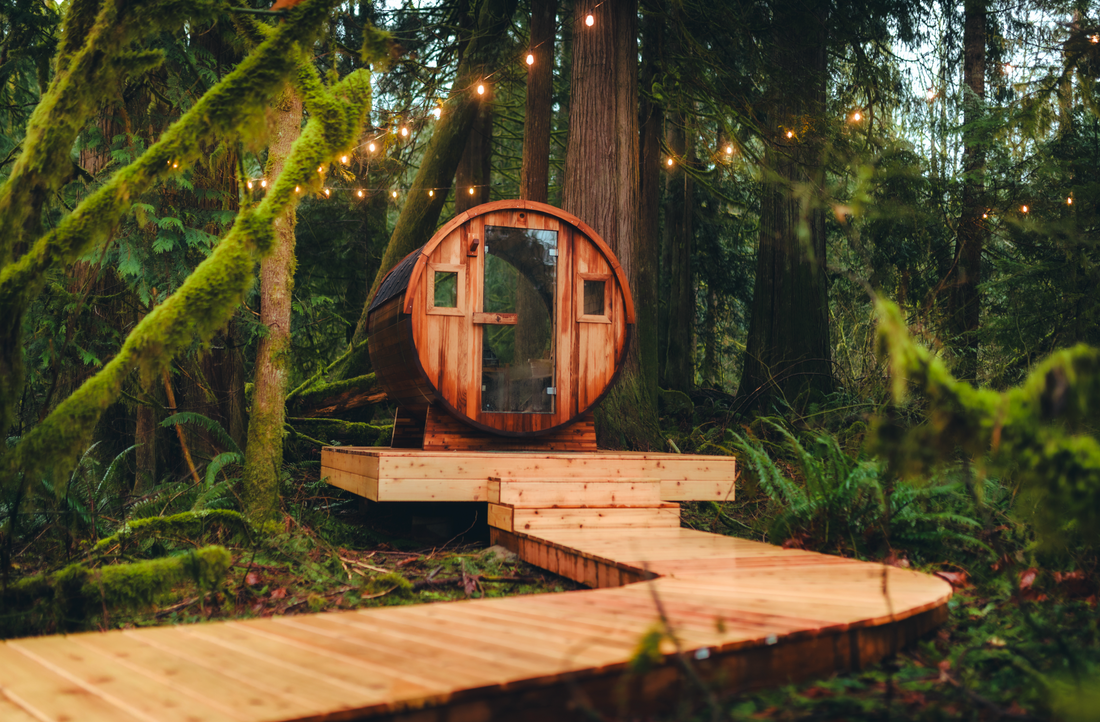 A Redwood Outdoors barrel sauna sits on a level gravel pad in a forest with open space on all sides.