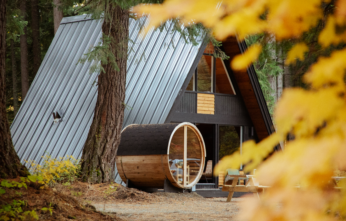 A Redwood Outdoors barrel sauna beside an A-frame cabin in a forest clearing, surrounded by autumn foliage.