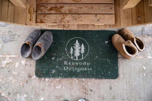 A Redwood Outdoors doormat in front of a sauna door, with a pair of boots on each side