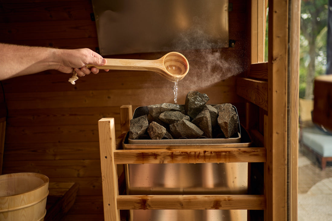 Water poured from a wooden ladle onto the hot stones of a Redwood Outdoors sauna heater, releasing a burst of steam.