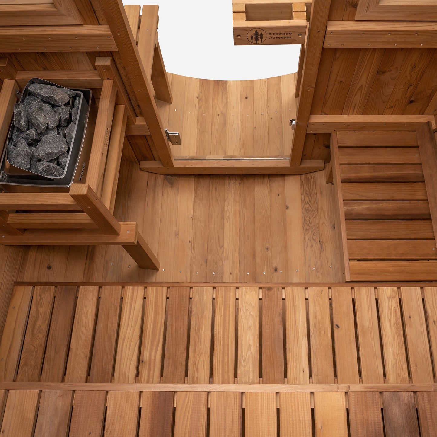 Redwood Outdoors 6 person Barrel Outdoor Extra-Wide Sauna With Porch made of hemlock, inside view from above, two benches with one on the side. A heater is visible on one side with a glass door and two windows. 
