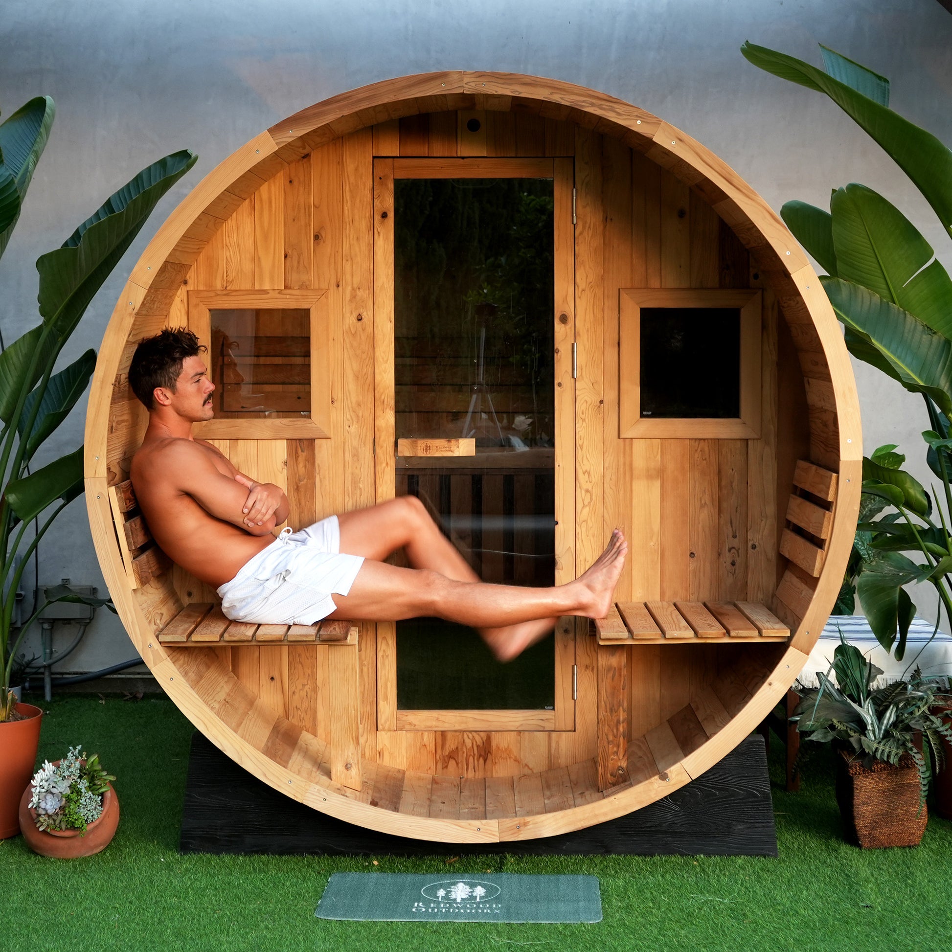 A person sits on the attached porch of a barrel sauna, with 2 glass windows and a full glass door