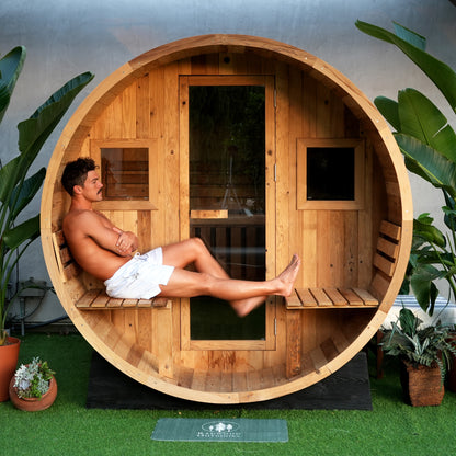 A person sits on the attached porch of a barrel sauna, with 2 glass windows and a full glass door