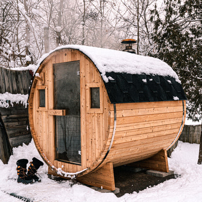 Redwood Outdoors 6 person Barrel Outdoor Sauna made of hemlock with a glass door and two windows, front outside lifestyle view in the snow. 