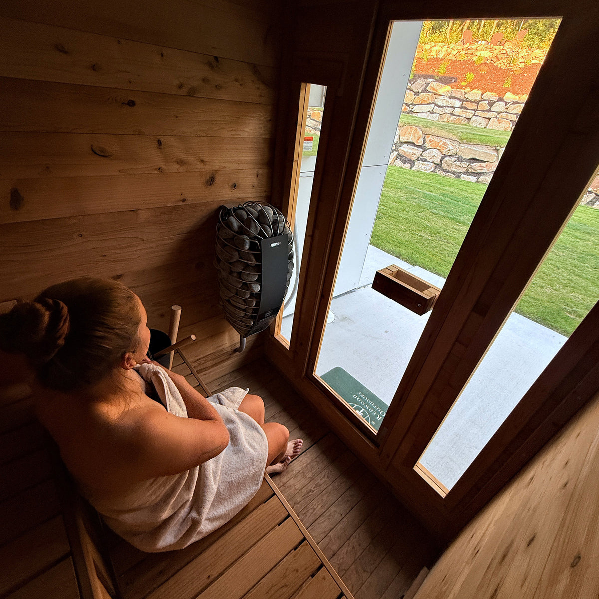 Redwood Outdoors Cabin 4 person outdoor sauna made of hemlock wood with two front windows. In a backyard with a view through the windows from the interior of the sauna. 