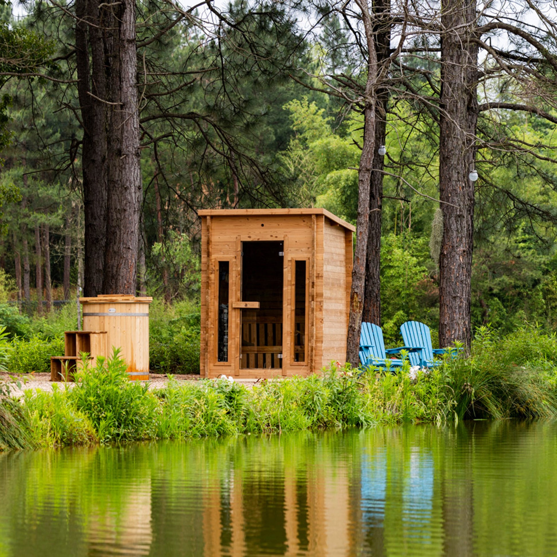 Redwood Outdoors Cabin 4 person outdoor sauna made of hemlock wood with two front windows. By a lake in the daylight, zoomed out view from the side of the sauna. 