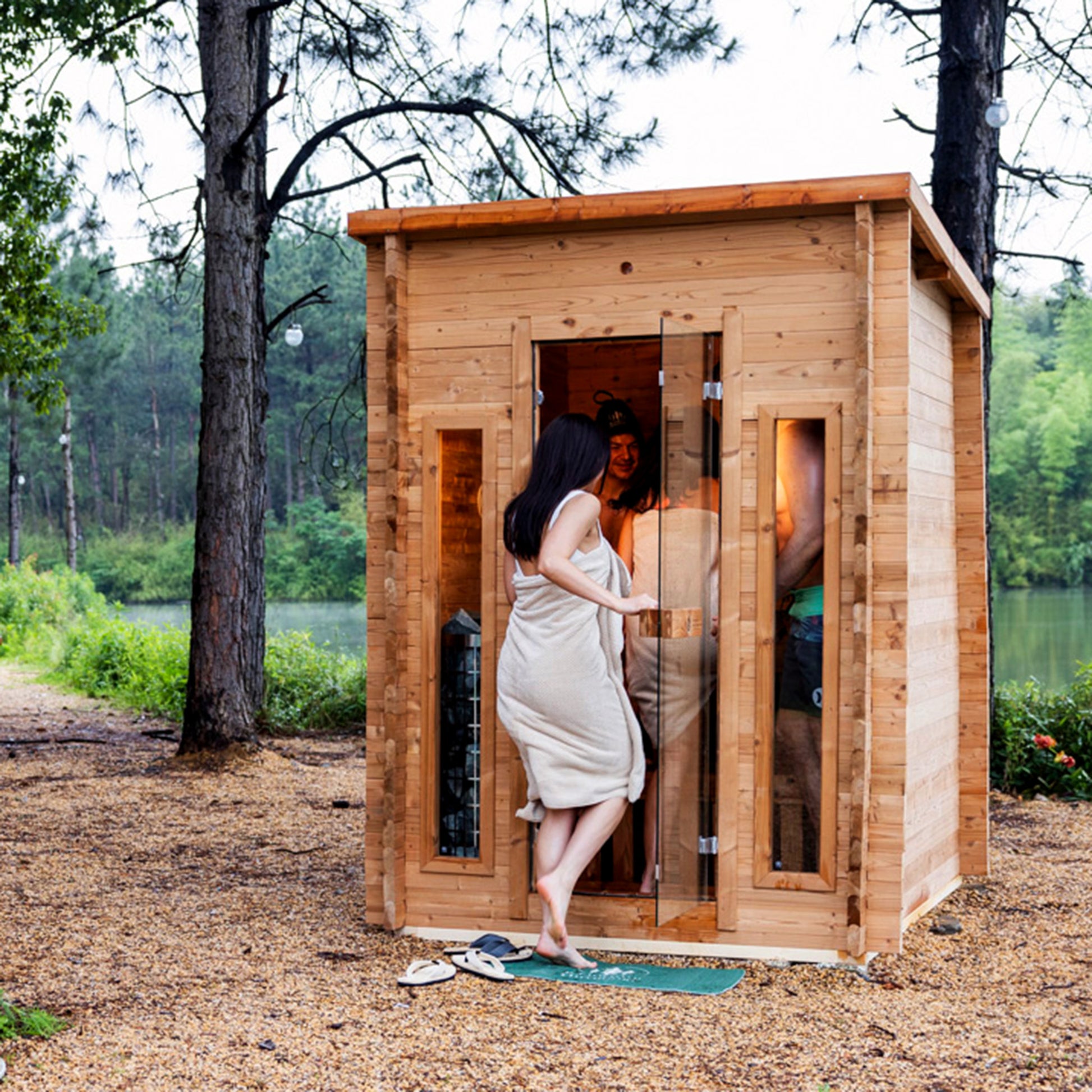 Redwood Outdoors Cabin 4 person outdoor sauna made of hemlock wood with two front windows. By a lake with people entering the sauna in the daylight. 