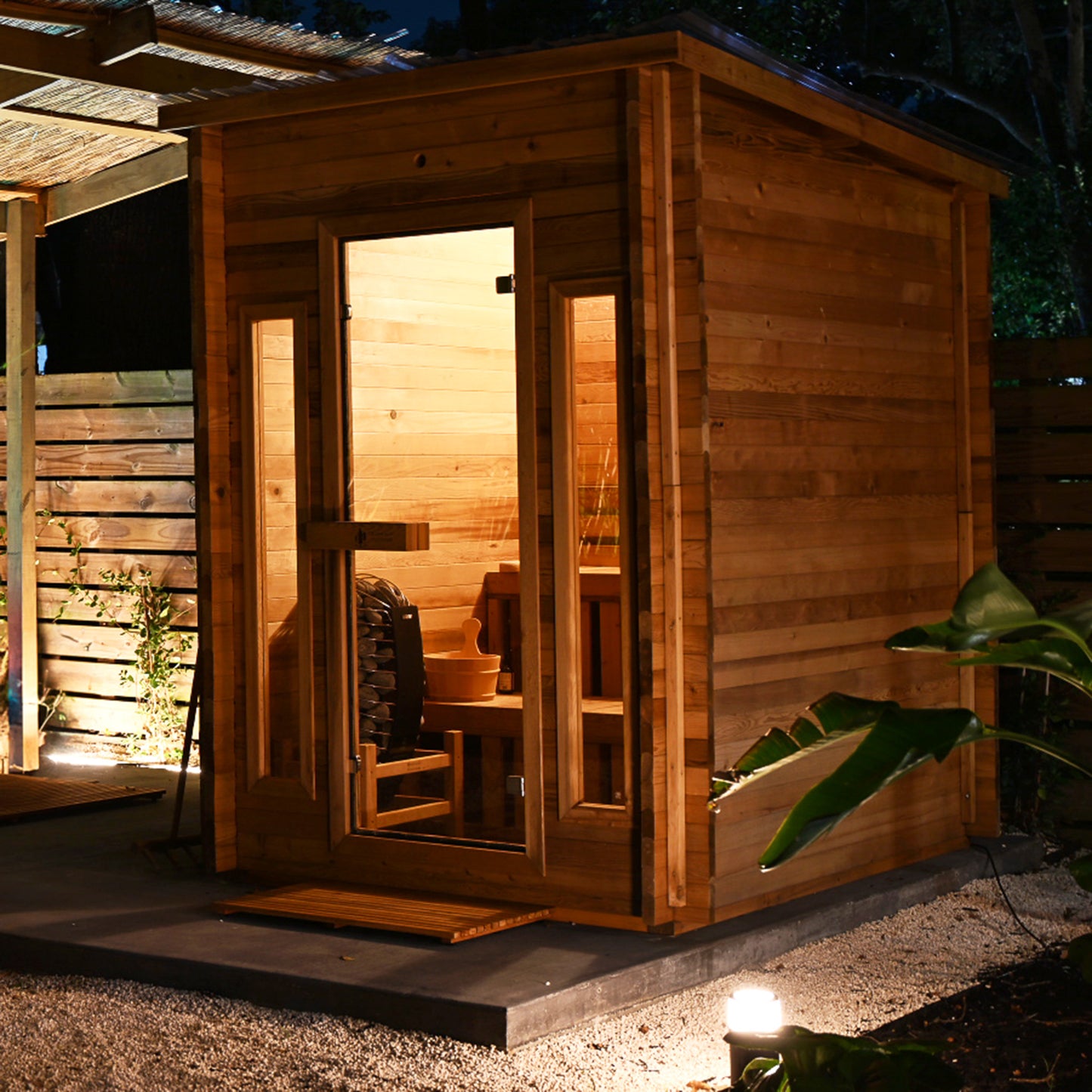 Redwood Outdoors Cabin 4 person outdoor sauna made of hemlock wood with two front windows. In a backyard at night with lights, side view. 