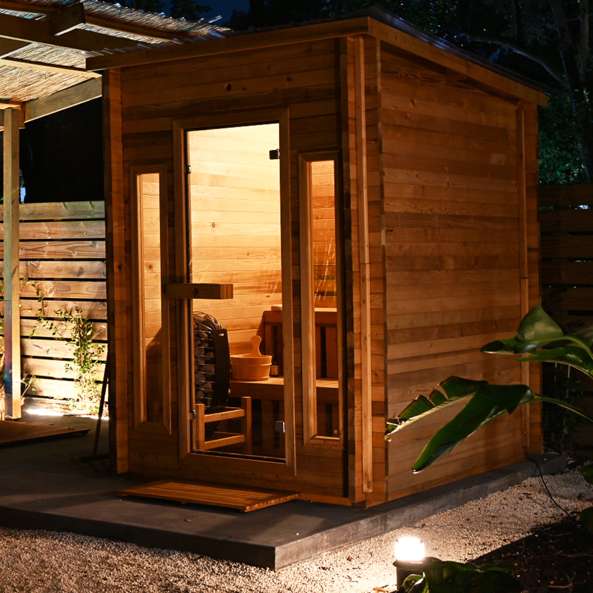 Redwood Outdoors Cabin 4 person outdoor sauna made of hemlock wood with two front windows. In a backyard at night with lights, side view. 