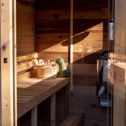 Redwood Outdoors Cove 3 person sauna, made of hemlock wood. A lifestyle view inside the sauna of an empty bench with light from the window shinning in with a wooden bucket and green hat placed on a wooden bench.