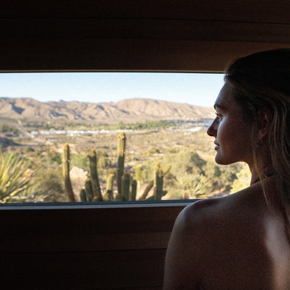Redwood Outdoors Cove 3 person sauna. A lifestyle view inside the sauna of a women looking out a large glass window onto a dessert landscape. 
