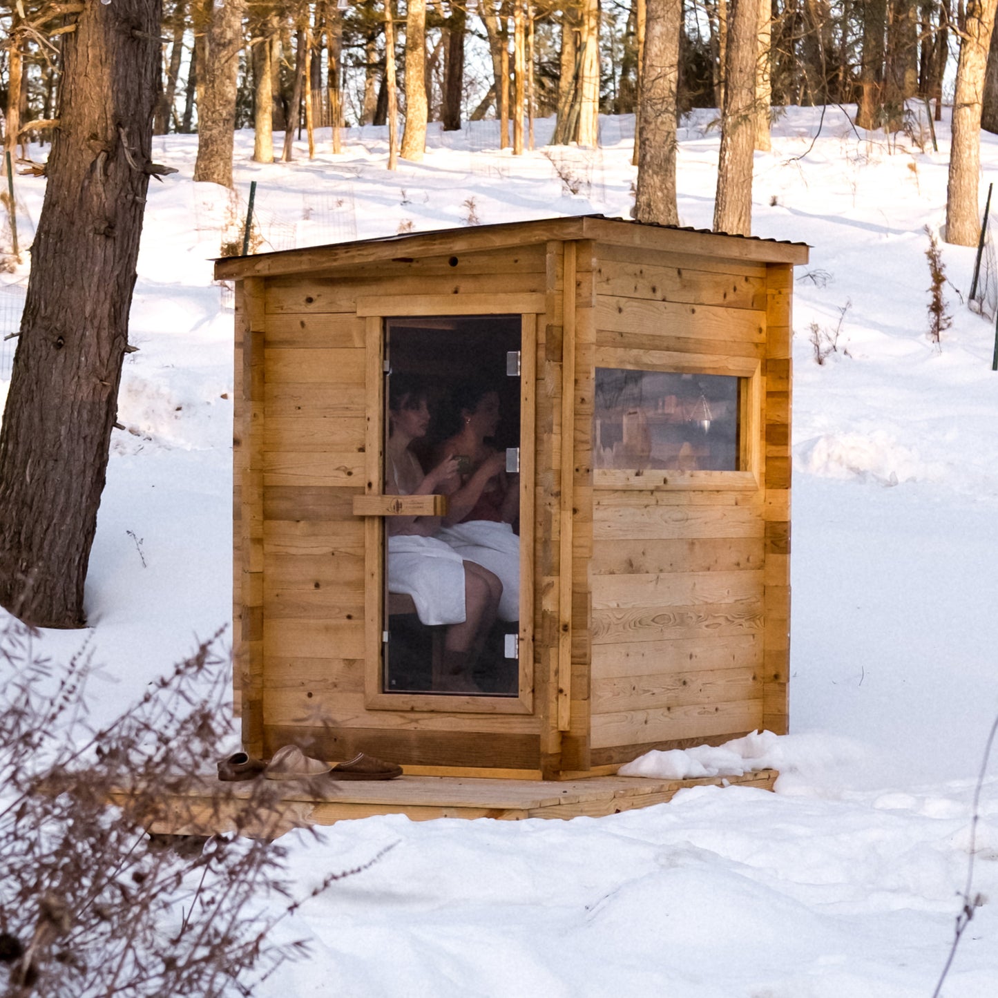 Redwood Outdoors Cove 3 person sauna, made of hemlock wood with glass door and large side glass window, view from the side of the sauna in the snow.