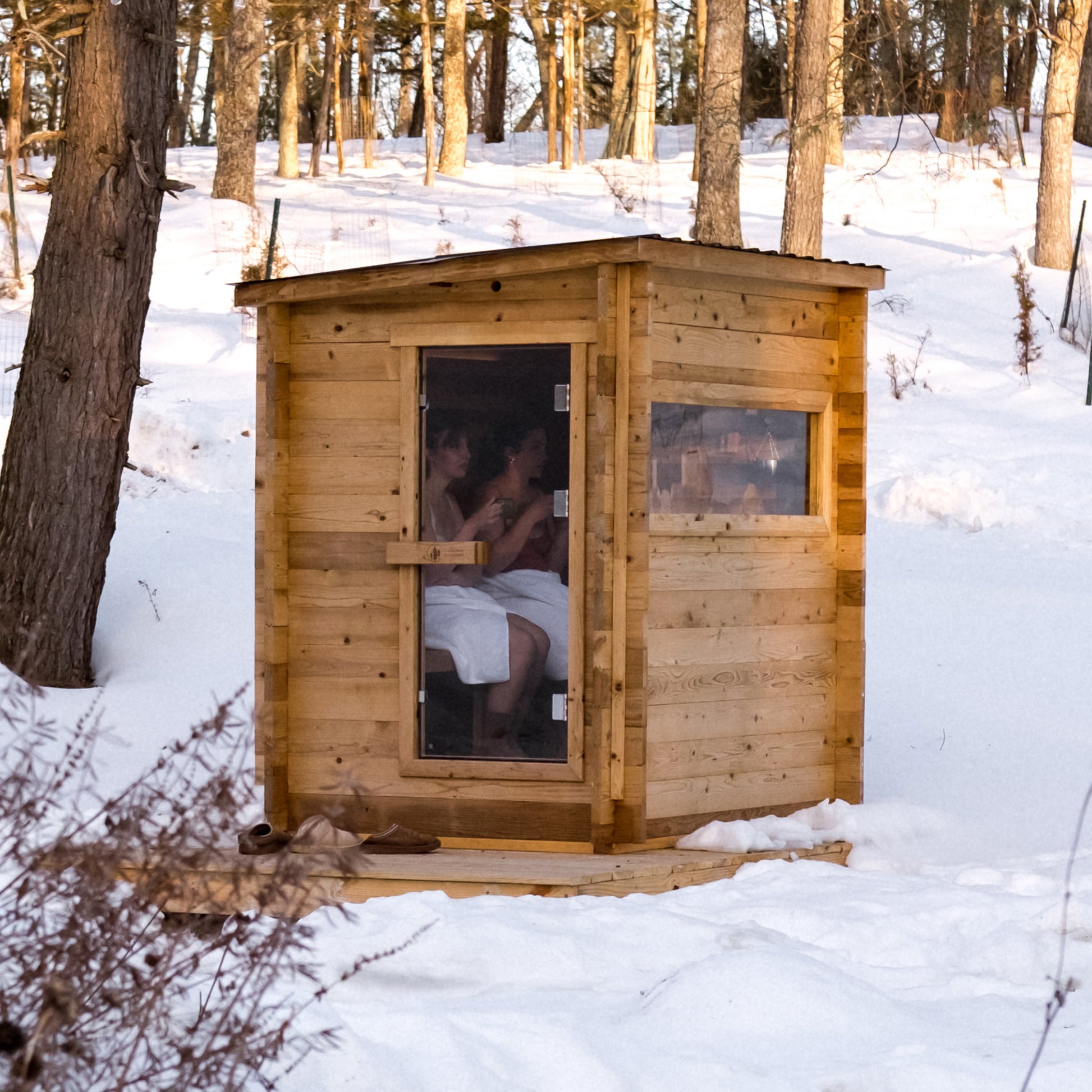 Redwood Outdoors Cove 3 person sauna, made of hemlock wood with glass door and large side glass window, view from the side of the sauna in the snow.