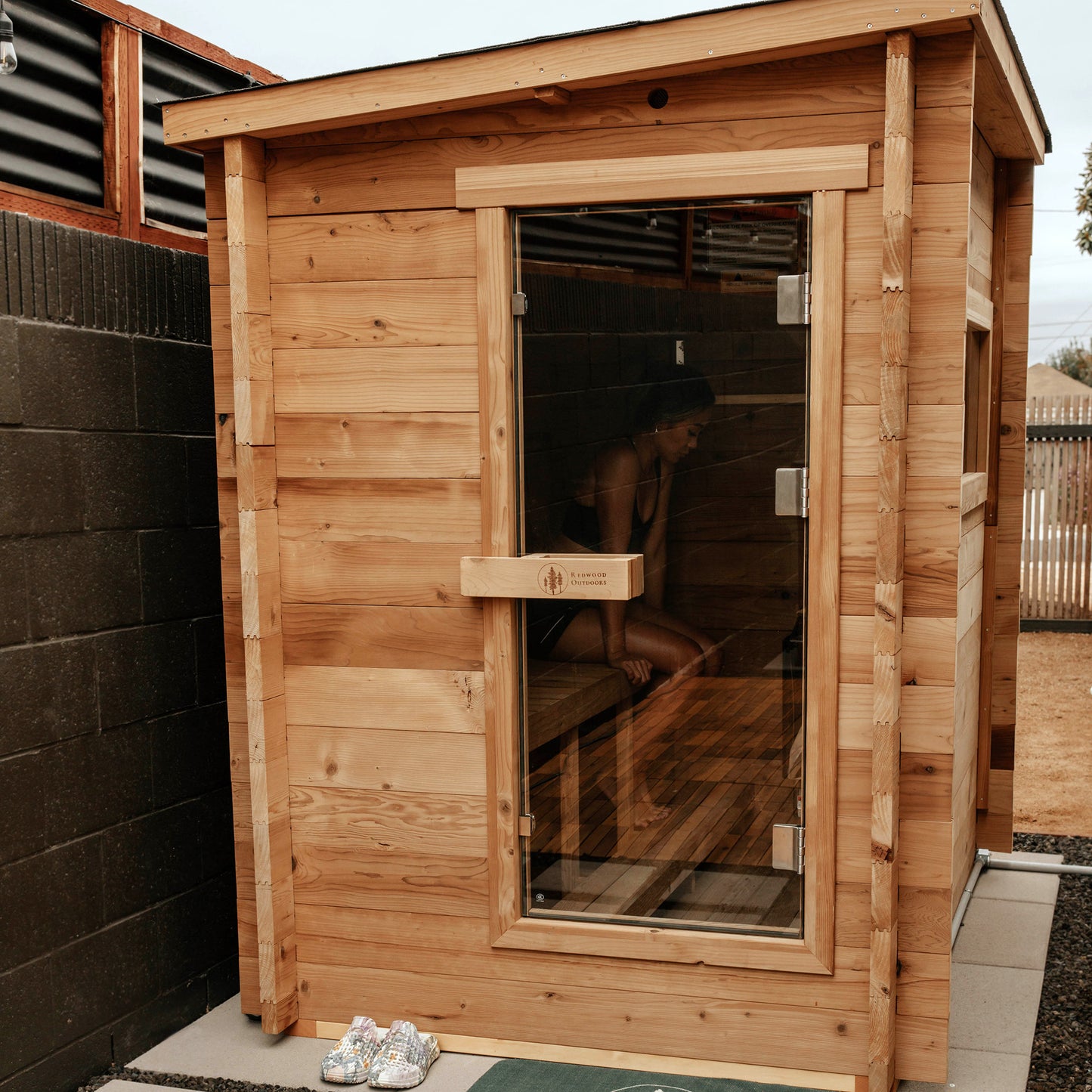 Redwood Outdoors Cove 3 person sauna, made of hemlock wood with glass door. A life style view of the sauna in a backyard with a women sitting inside.