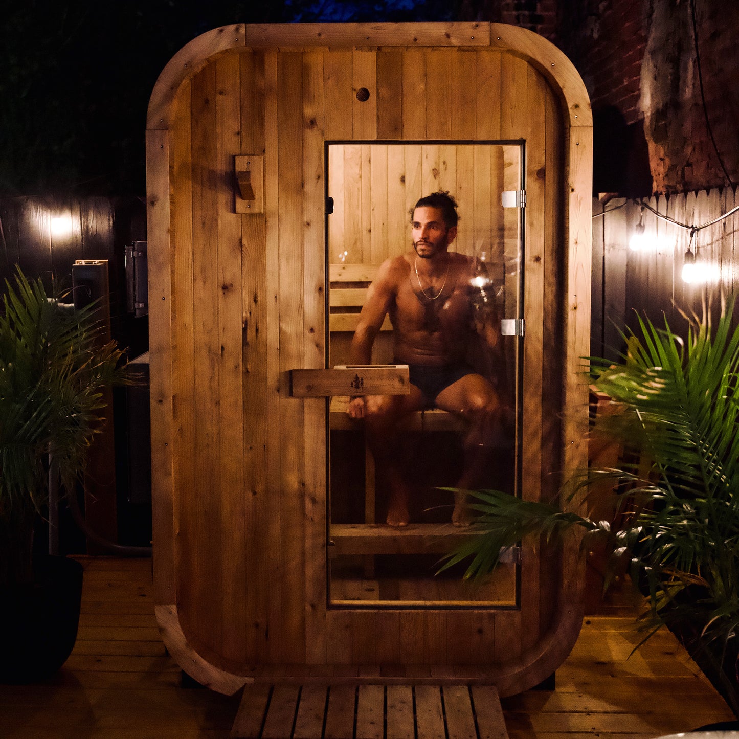 Redwood Outdoors Duo 2 person outdoor sauna, made of hemlock wood with a glass door with a person sitting inside at night