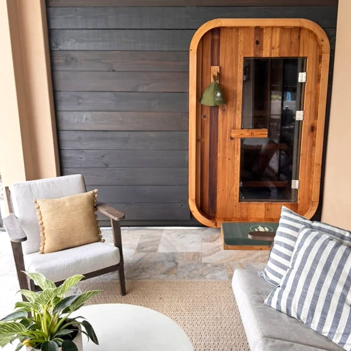 Redwood Outdoors Duo 2 person outdoor sauna, made of hemlock wood with a glass door outside on a porch with patio furniture in front