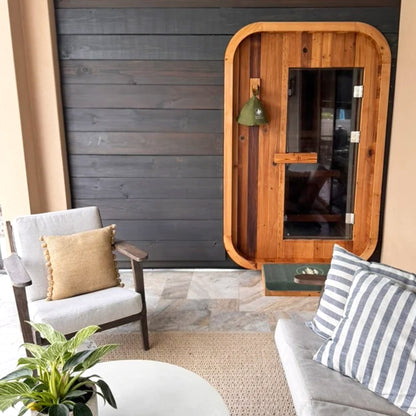 Redwood Outdoors Duo 2 person outdoor sauna, made of hemlock wood with a glass door outside on a porch with patio furniture in front
