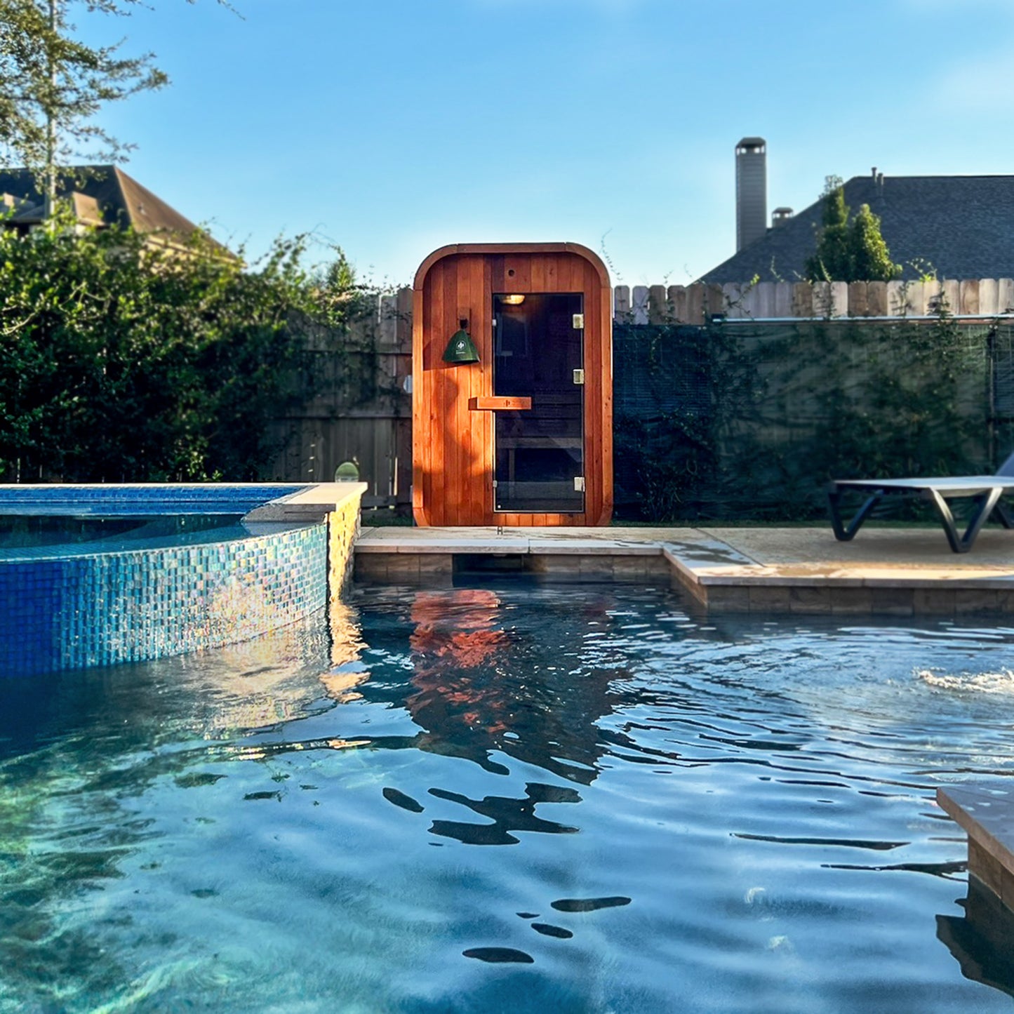 Redwood Outdoors Duo 2 person outdoor sauna, made of hemlock wood with a glass door on the other side of a pool. 