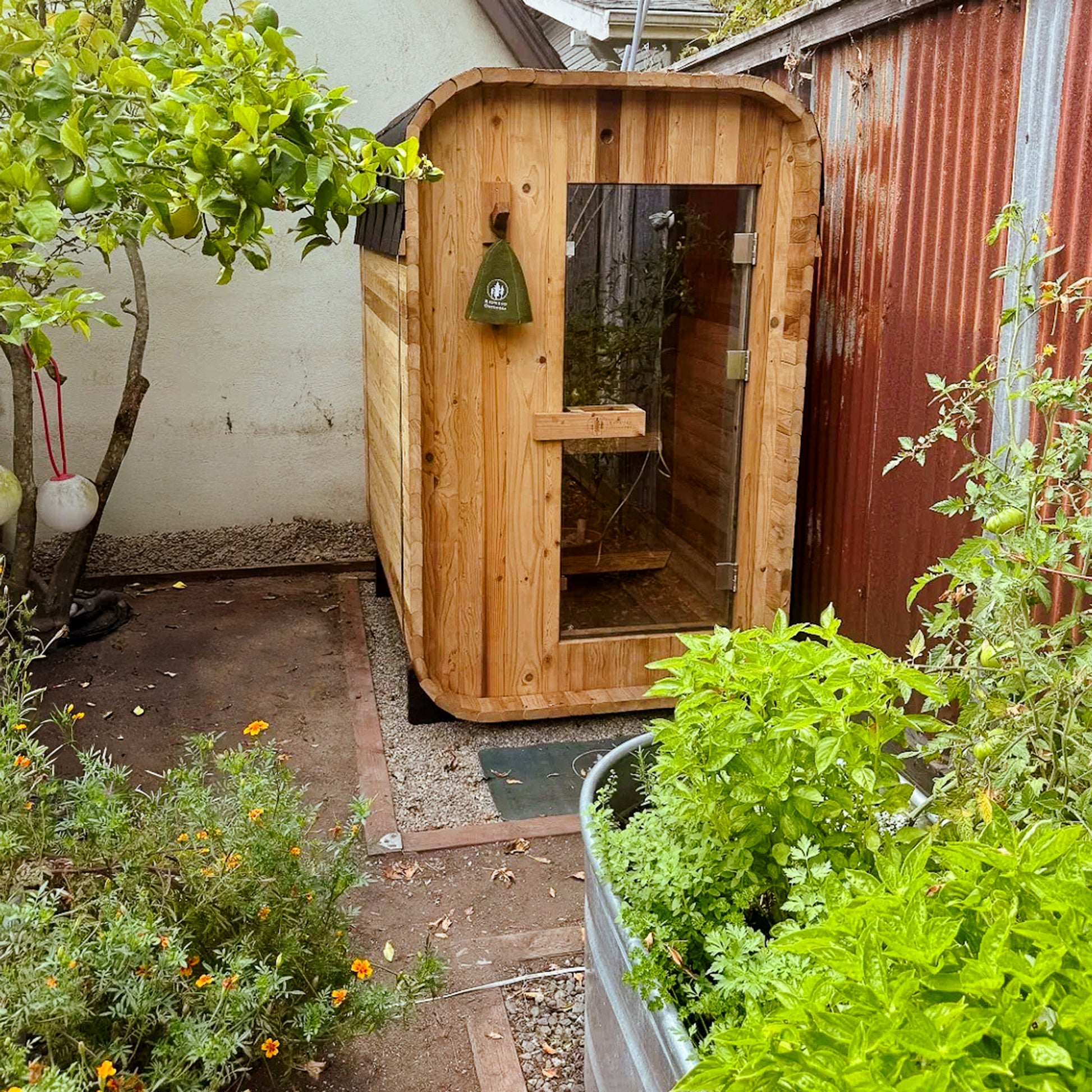 Redwood Outdoors Duo 2 person outdoor sauna, made of hemlock wood with a glass door outside on a porch in a garden surrounded by plants