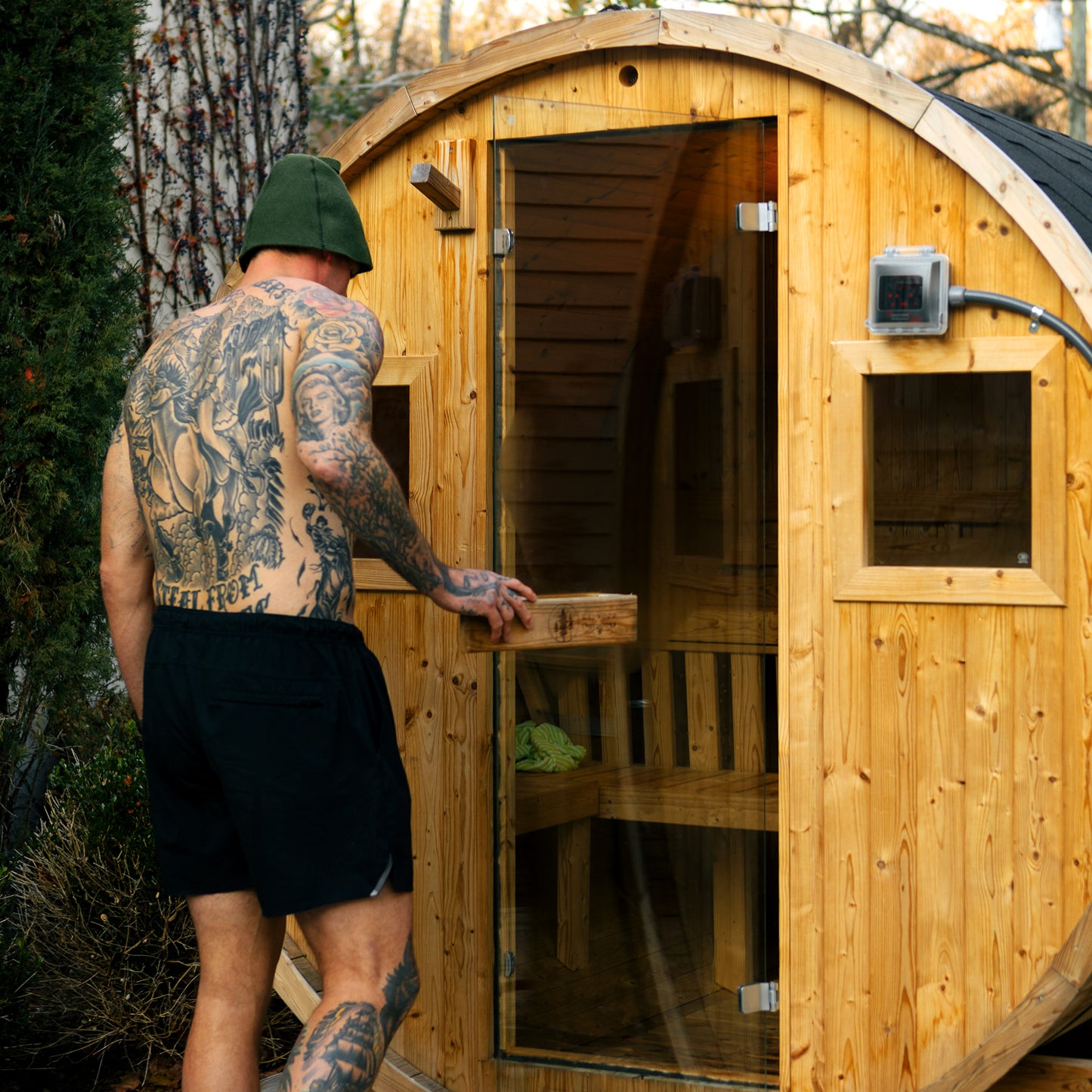 Redwood Outdoors Extra-Wide 6 person Barrel Sauna lifestyle front angle view in a backyard with plants behind and a person entering the sauna. 