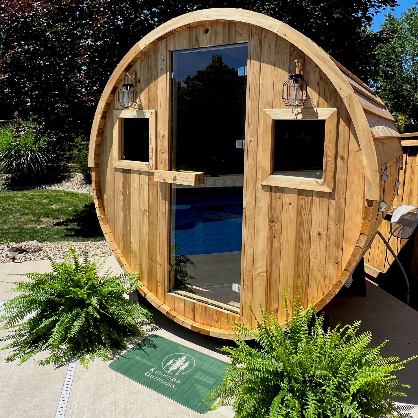 Redwood Outdoors Extra-Wide 6 person Barrel Sauna lifestyle front angle view in a backyard with plants in front. 
