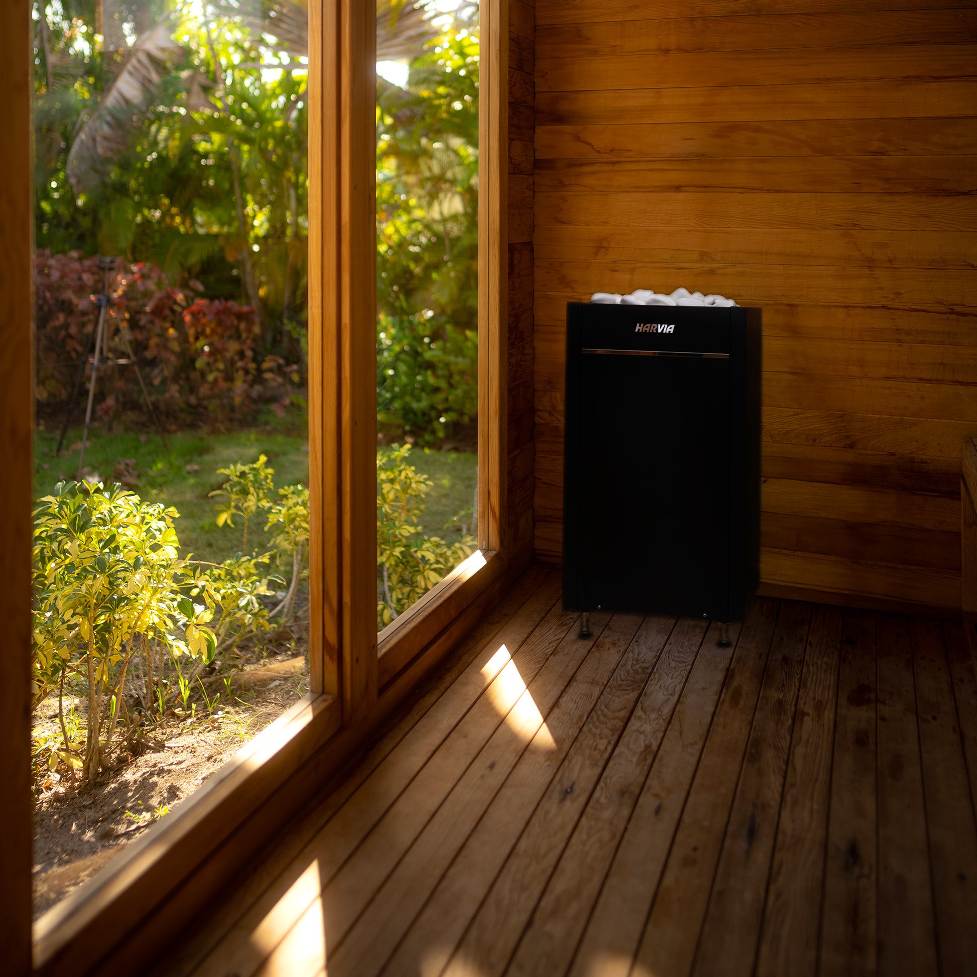 Redwood Outdoors Garden 8 person sauna. View from inside of the sauna looking out of the large glass window onto a garden with a heater in the corner. 