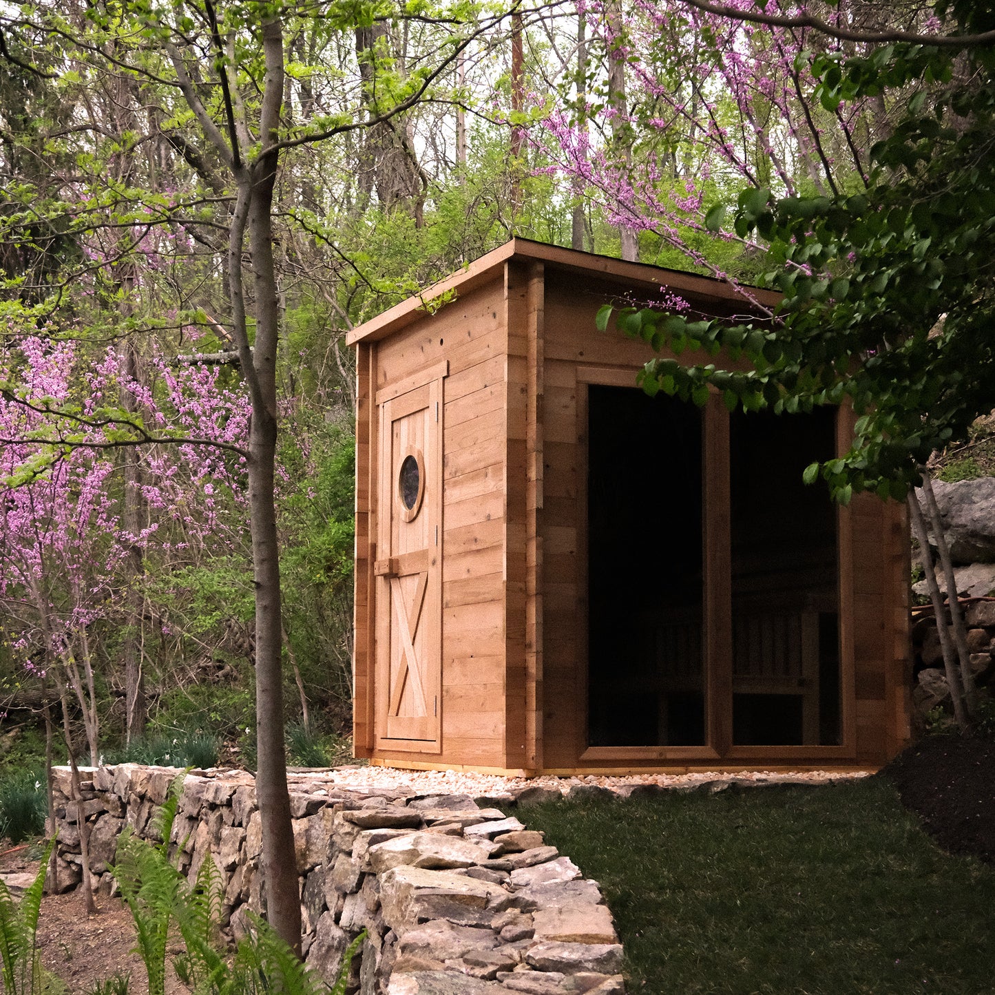 Redwood Outdoors Garden 8 person sauna in a back yard with trees and flowers. A view from the side showing the wood door and large glass side window. 