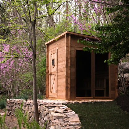 Redwood Outdoors Garden 8 person sauna in a back yard with trees and flowers. A view from the side showing the wood door and large glass side window. 