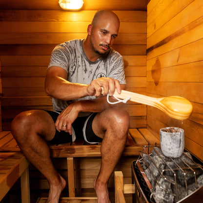 Redwood Outdoors Haven 3 person indoor sauna, made of hemlock wood interior lifestyle view of a person sitting in the sauna pouring water over the rocks 