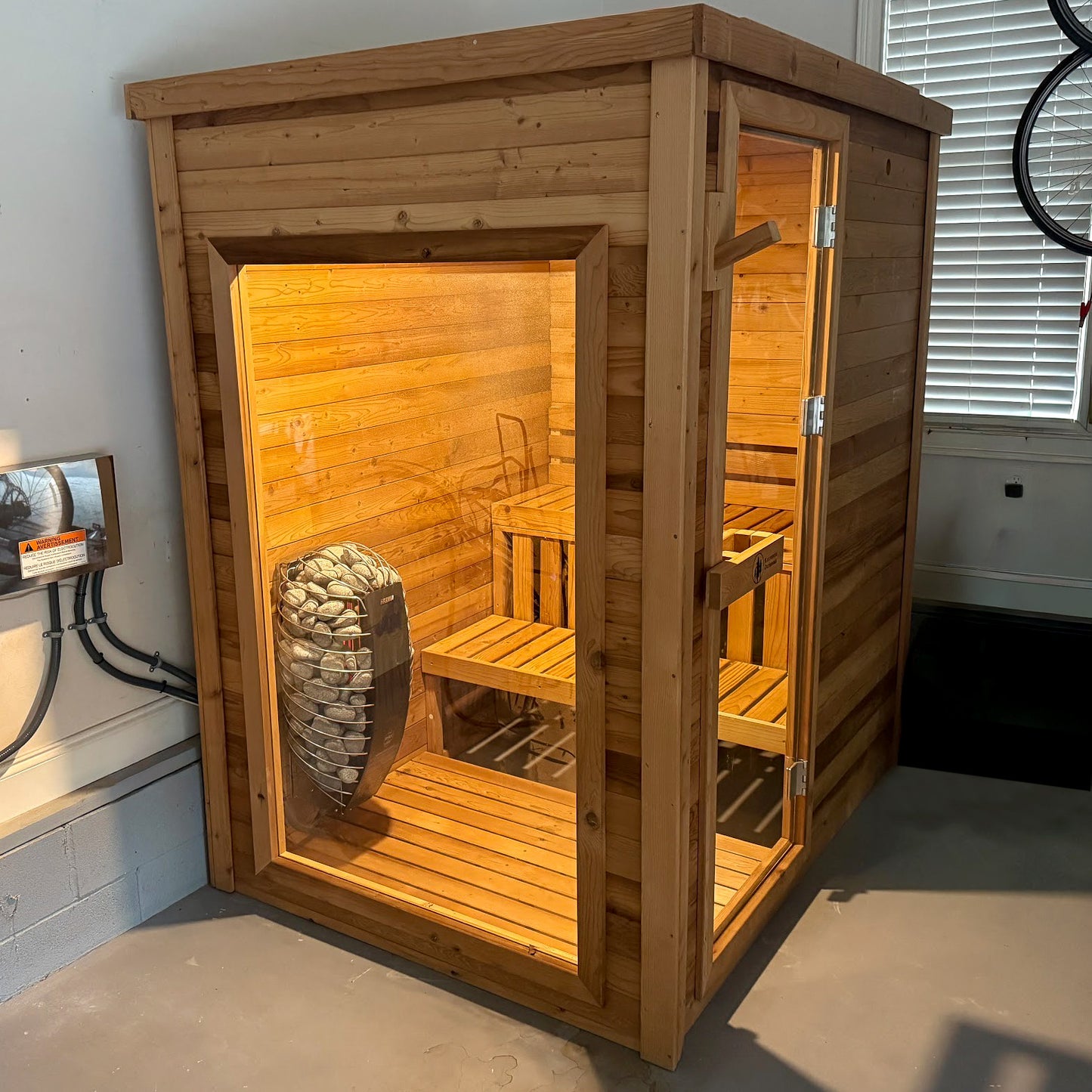 Redwood Outdoors Sanctuary 4 person indoor sauna, made of hemlock wood with a glass door and window exterior front view in a garage