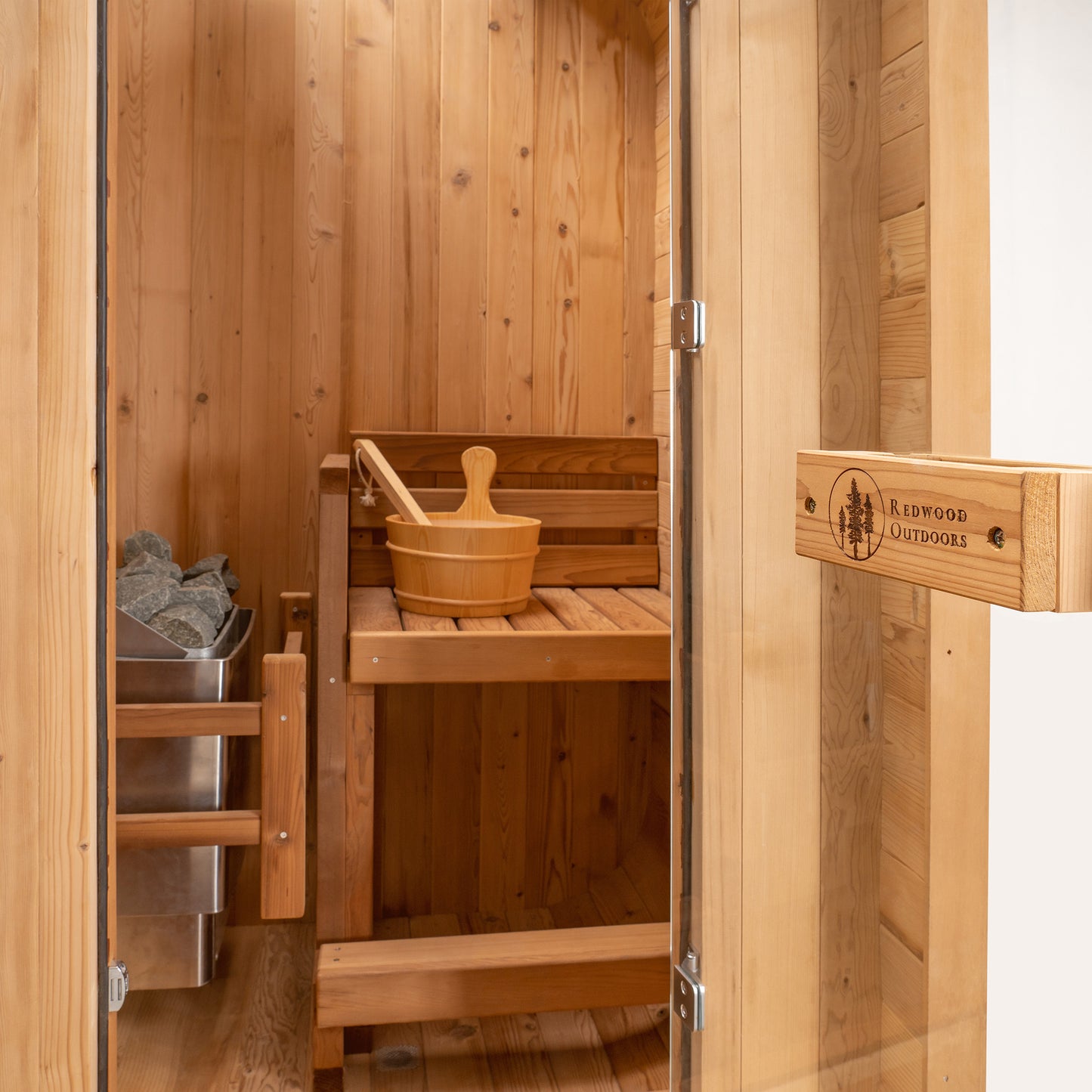 Redwood Outdoors Solo 1 person outdoor sauna, made of hemlock wood with a glass door open to the inside of the sauna with a bench, heater and bucket in view. 