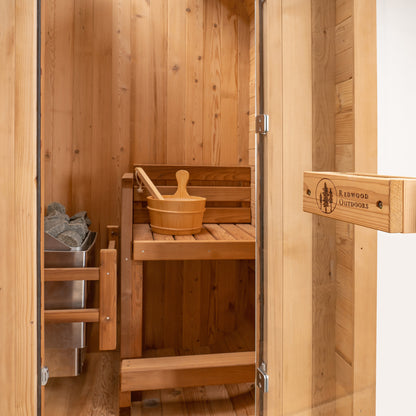 Redwood Outdoors Solo 1 person outdoor sauna, made of hemlock wood with a glass door open to the inside of the sauna with a bench, heater and bucket in view. 