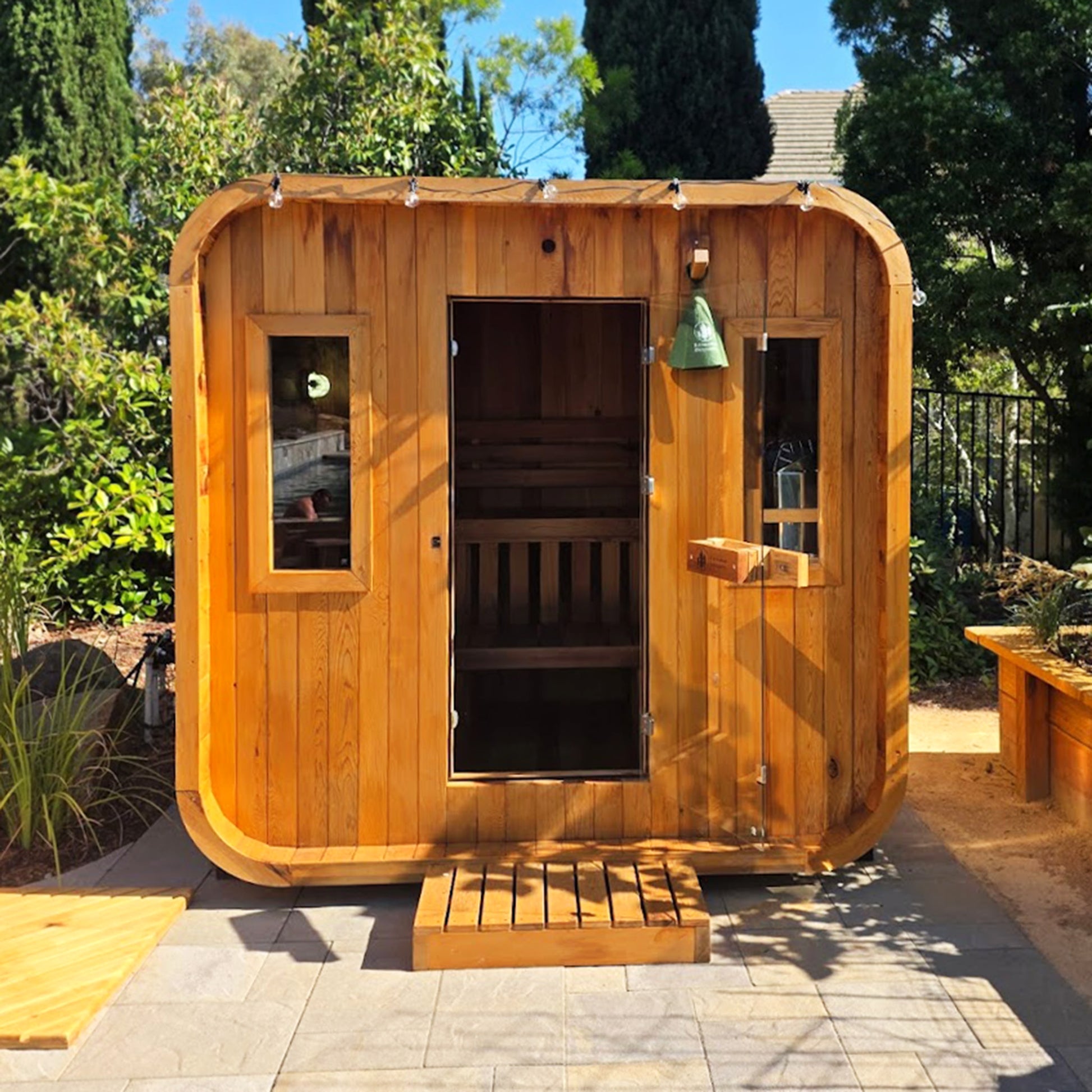 Redwood Outdoors Summit 6 person outdoor sauna, made of hemlock wood with a glass door and two large windows, lifestyle exterior view of the sauna in a garden. 
