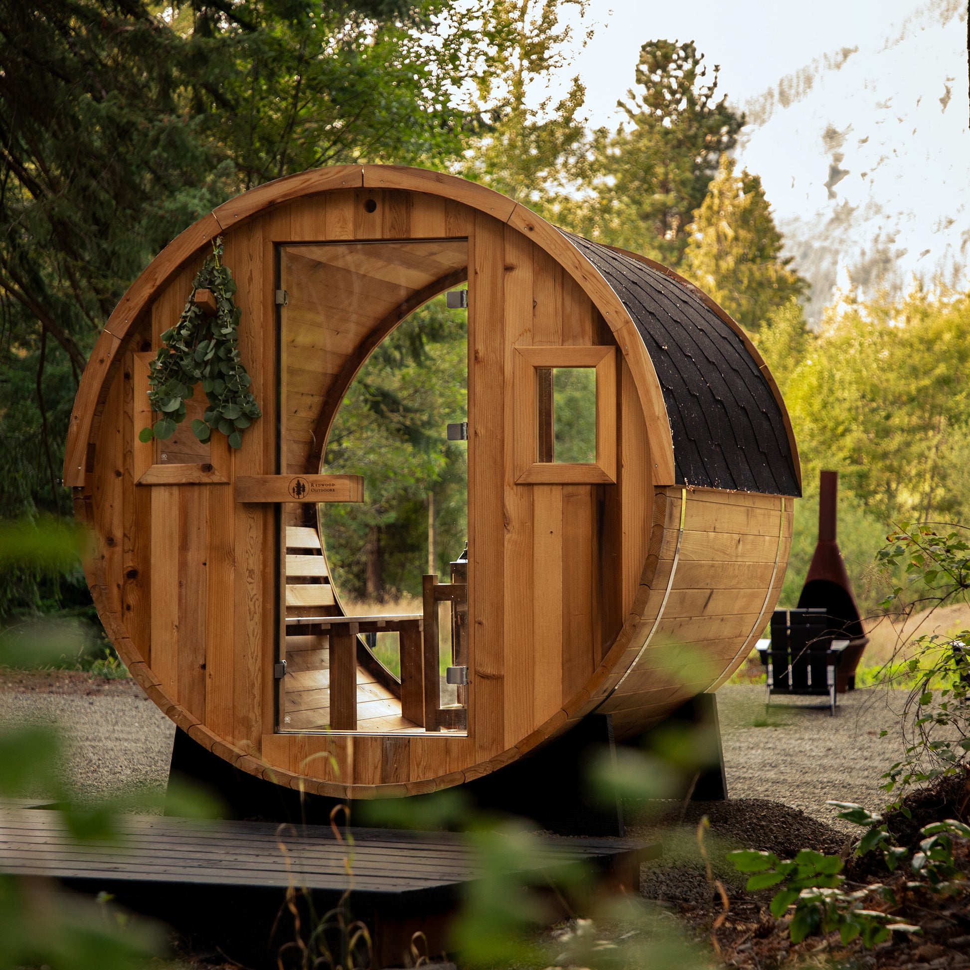 Redwood Outdoors Vista 6 person outdoor sauna, made of hemlock wood with glass door and large rear window, view from a front side angle with woods in the background. 