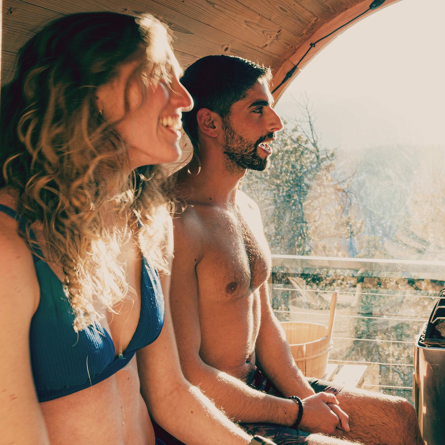 Redwood Outdoors Vista 6 person outdoor sauna, made of hemlock wood with glass door and large rear window, view from inside the sauna with two people sitting on a bench with trees in the background. 