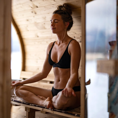 Redwood Outdoors Vista 6 person outdoor sauna, made of hemlock wood with glass door and large rear window, view from inside the sauna of a women sitting on the bench. 